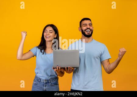 Concetto di grande successo. Felice coppia araba che tiene il laptop, acclamando e alzando le mani, celebrando la vittoria, sfondo giallo Foto Stock