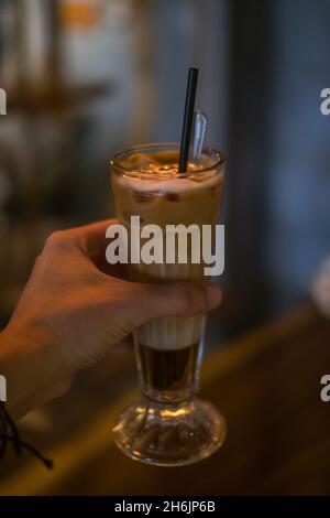 Foto ravvicinata della tazza di vetro con latte di ghiaccio in un bar. Mano che tiene il bicchiere con cappuccino al caramello ghiacciato con la cannuccia nel ristorante. Foto Stock