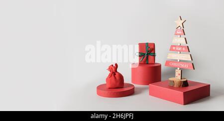 Banner natalizio con albero di Natale, regalo rosso e borsa regalo sul podio su sfondo grigio. Biglietto d'auguri moderno di Natale con spazio per la copia. Concetto di sus naturale Foto Stock