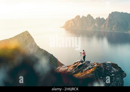 Fotografo con treppiede sulla montagna Husfjellet che guarda il tramonto sulle cime taglienti del monte Okshunnan (denti del diavolo, Senja, Norvegia, Scandinavia, Europa Foto Stock
