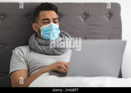 Malato giovane arabo uomo che indossa maschera medica lavoro con laptop in letto Foto Stock