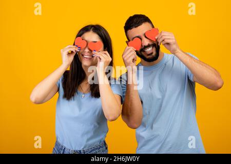 Accecato dall'amore. Giovane coppia araba romantica con cuori di carta rossa che coprono gli occhi, posando su sfondo giallo Foto Stock