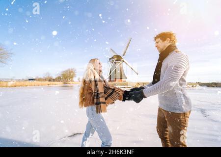 Coppia amorevole divertirsi sul ghiaccio nel tipico paesaggio olandese con mulino a vento. Pattinaggio su ghiaccio uomo e donna all'aperto in giornata di neve soleggiata. Romantica data attiva sul canale ghiacciato nella vigilia di Natale d'inverno. Foto Stock