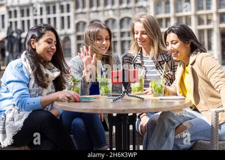 Anversa, Belgio, 21 maggio 2021, quattro turisti o studenti di razza mista seduti fuori nel centro storico della città in una caffetteria terrazza con una videochiamata utilizzando il telefono cellulare. Foto di alta qualità Foto Stock