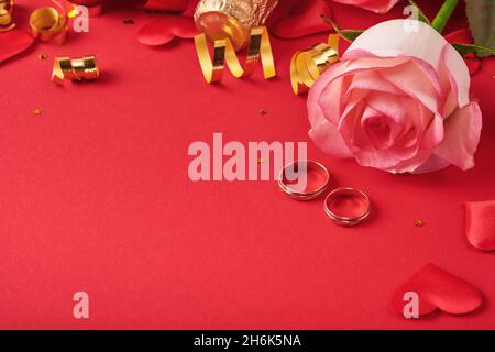 Rosa rose fiori, anelli di nozze, champagne, regalo, nastri dorati e confetti cuori su sfondo rosso. Vista dall'alto con spazio per i messaggi di saluto. San Valentino Foto Stock