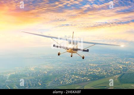 L'aereo leggero vola nelle nuvole del cielo mattutino all'alba sopra la città, vista posteriore Foto Stock