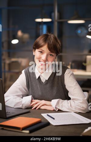Ritratto di giovane donna d'affari sorridente alla macchina fotografica mentre si siede al suo posto di lavoro in ufficio Foto Stock