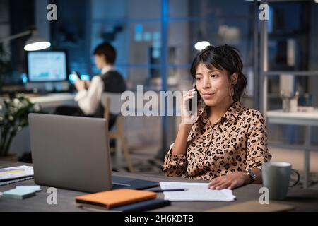 Donna d'affari matura seduta sul posto di lavoro con documenti che guardano lo schermo del laptop e parlano al telefono mentre lavora in ufficio la sera Foto Stock