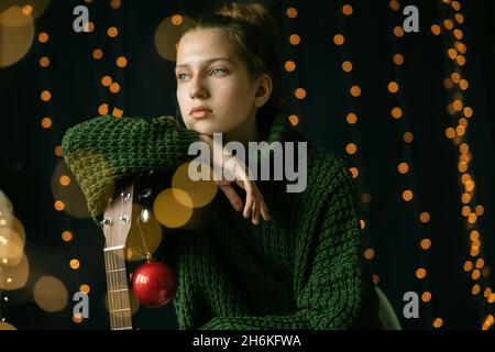 Una bella ragazza teenager guarda da sogno a lato, appoggiata sul collo di una chitarra. Decorazioni natalizie e illuminazione a casa. Stile di vita, orizzonte relax Foto Stock