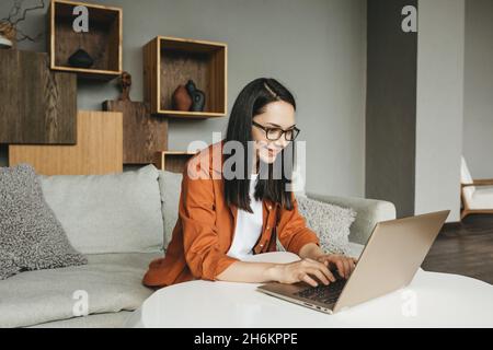 Una bella giovane donna europea che usa un computer portatile per lavorare a distanza da casa. Una donna d'affari in abiti casual conduce una videochiamata, si impegna in e-mail, comunica online con i colleghi, invia e risponde alle e-mail, sviluppa software mentre si siede a casa sul divano. Una donna intelligente lavora da remoto Foto Stock