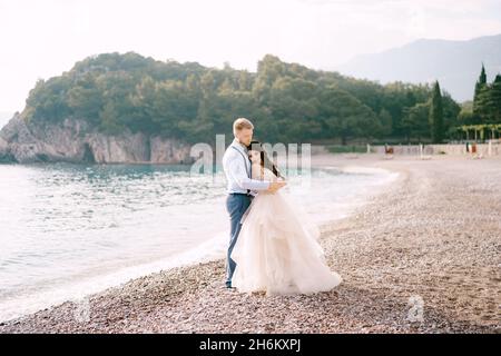 La sposa e lo sposo si erigono sulla spiaggia di ciottoli e abbracciano, lo sposo abbraccia le spalle della sposa Foto Stock