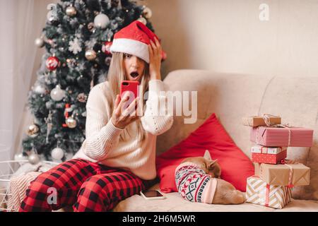 Sorpresa giovane donna e cane in maglione prende selfie ritratto sullo smartphone durante le vacanze invernali, godere il tempo di Natale a casa. Confezione regalo e. Foto Stock