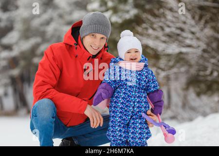 Sorridente bambina che si diverte con il padre nel parco invernale. Carino bambino di due anni con il genitore che gioca il giocattolo del creatore delle palle di neve. Concetto di famiglia felice e. Foto Stock