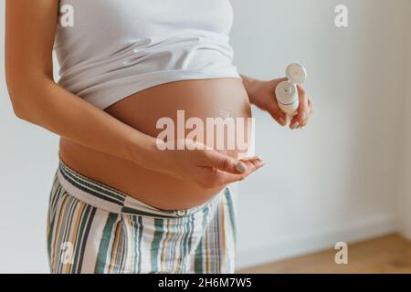 Donna bionda incinta che applica la crema per il ventre per impedire i segni di stiramento. Futura madre tenuta in tubo con lozione per la cura della pelle. Foto Stock