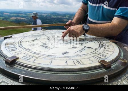 Vista ravvicinata del quadrante panoramico sulla cima con un camminatore maschile che punta sulla mappa, mentre i visitatori si trovano al di là sotto la luce del giorno, Malvern Hills, Inghilterra Foto Stock