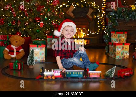 Il ragazzo piccolo felice in cappello di Babbo Natale gioca con la locomotiva a casa mentre si siede vicino all'albero di Natale. Foto Stock