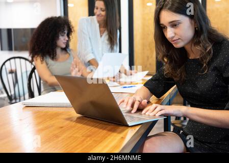 Giovane donna che lavora sul laptop con donne d'affari che parlano in background Foto Stock