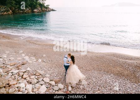 La sposa e lo sposo si levano in piedi sulla spiaggia ciottolata e abbracciano Foto Stock