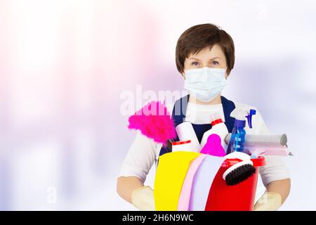 Il concetto di servizio di pulizia. Un secchio di prodotti per la pulizia in mani con guanti di gomma donna in uniforme blu Foto Stock