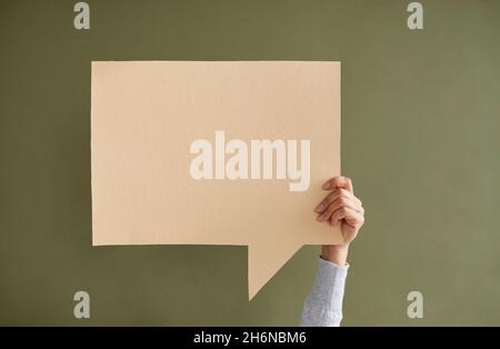 Giovane donna anonima che tiene una bolla vuota di discorso quadrato beige in mano su sfondo verde. Foto Stock
