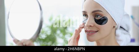 Giovane donna con macchie nere sotto gli occhi e con asciugamano sulla testa guardando nello specchio a casa Foto Stock