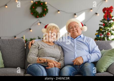 Sorridente coppia matura in pensione a Natale Babbo Natale cappelli seduta, abbracciando e guardando la macchina fotografica Foto Stock