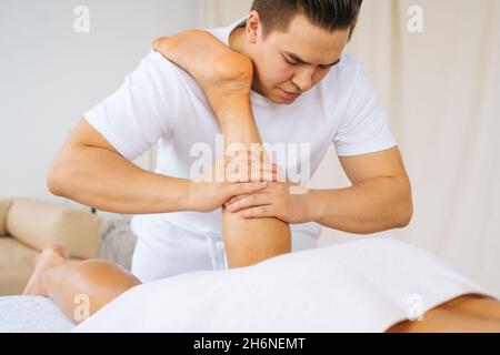 Primo piano viso maschile professionale con mani forti massaggiando i muscoli del polpaccio sulla gamba inferiore dell'atleta muscolare dopo l'allenamento sportivo fisico Foto Stock