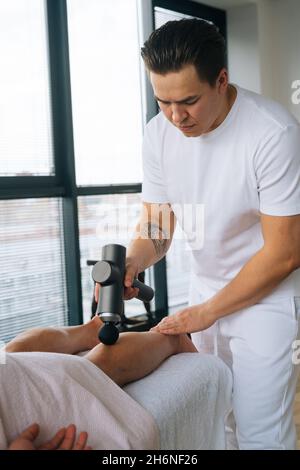 Colpo verticale del massaggiatore maschile professionale massaggiante i muscoli del polpaccio delle gambe utilizzando la pistola da massaggio percussione strumento di atleta muscolare uomo. Foto Stock