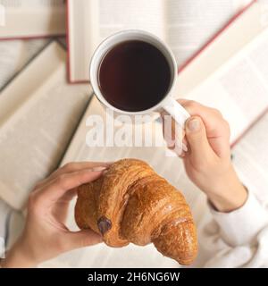 Mani di una giovane donna che mangia un croissant con caffè sullo sfondo di un mucchio di libri. Studente che mangia la colazione, concetto di studio. Foto Stock