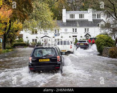 I veicoli percorrono una strada allagata a Waterhead, Ambleside, Lake District, Regno Unito, dopo piogge torrenziali che hanno portato il lago Windermere a raggiungere livelli molto elevati Foto Stock