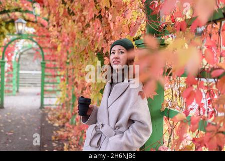 Donna in cappotto di lana e cappello verde lavorato a maglia a piedi e bere caffè da tazza riutilizzabile al parco autunnale Foto Stock