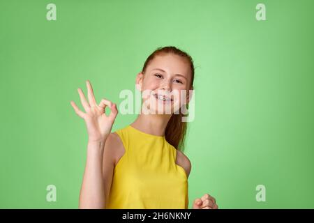Giovane ragazza rossa che mostra il gesto OK su sfondo verde. Foto Stock