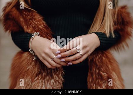 Mani femminili con manicure rosa, braccialetto argento e anelli. Figura femminile ritagliata con giubbotto marrone in pelliccia Foto Stock