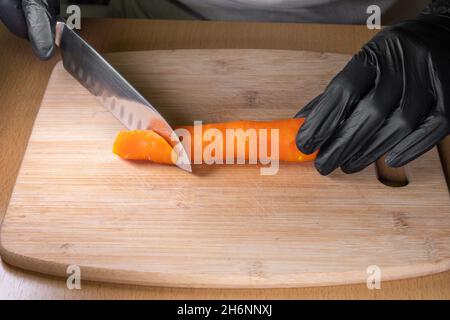 Le mani dello chef in guanti neri che tengono un coltello e tagliano le carote pelate su un asse di legno da cucina, primo piano. Il processo di preparazione di un'insalata. Naturale Foto Stock