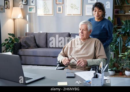 Donna matura che si prende cura dell'anziano a casa, lo porta su una sedia a rotelle al tavolo con il computer Foto Stock
