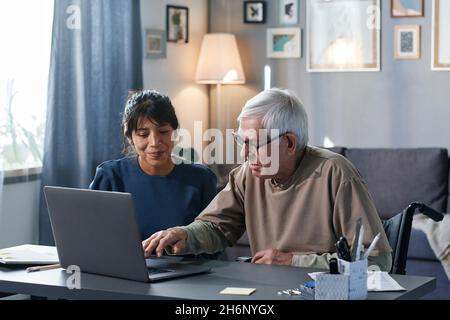 Uomo anziano imparando ad usare il computer con l'aiuto della donna mentre si siedono al tavolo insieme nella stanza Foto Stock