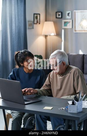 L'uomo anziano imparando fare lo shopping in linea con l'aiuto del volontario che si siede al tavolo nella stanza Foto Stock