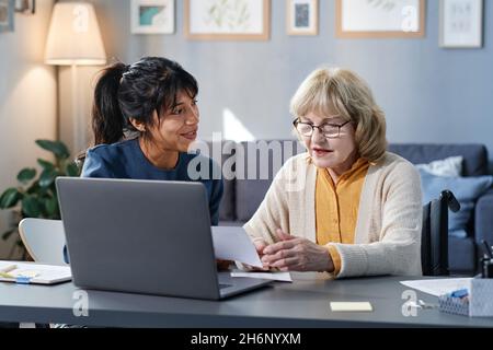 Donna anziana che studia al tavolo con il laptop, e volontario che la aiuta nel suo studio domestico Foto Stock