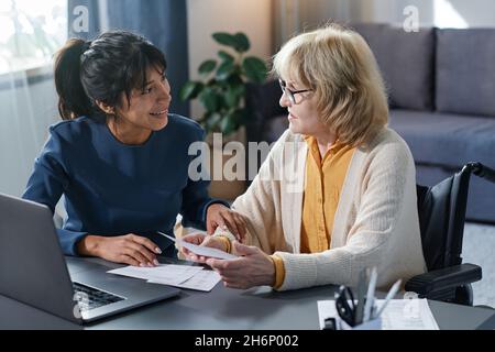 Sorridente volontario che aiuta la donna anziana a pagare le fatture in linea mentre si siedono al tavolo nella stanza Foto Stock