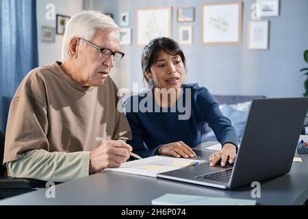 Uomo anziano seduto al tavolo di fronte al portatile e studiare online insieme al volontario a casa Foto Stock