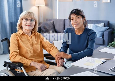 Ritratto della donna anziana con sedia a rotelle che guarda la fotocamera mentre si siede nella stanza insieme all'operatore domestico Foto Stock