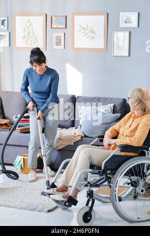 Donna anziana che usa la sedia a rotelle che parla con la governante che aspira il pavimento nella stanza Foto Stock