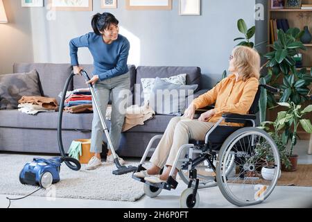 Cameriera che usa l'aspirapolvere per pulire la moquette nella stanza, aiutava la donna anziana che usa la sedia a rotelle Foto Stock