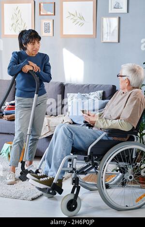 Uomo anziano che usa la sedia a rotelle che parla con la cameriera che pulisce la stanza, che le dà le istruzioni prima di lavorare Foto Stock