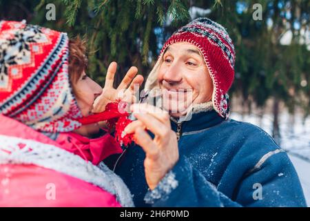Coppia di famiglie anziane che camminano nella foresta invernale. L'uomo e la donna godono di attività invernali. L'uomo stringe il cappello di sua moglie divertendosi. Attenzione Foto Stock