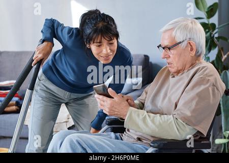 Uomo anziano in occhiali utilizzando la sedia a rotelle di apprendimento per utilizzare il telefono cellulare, egli chiedendo aiuto alla sua cameriera Foto Stock