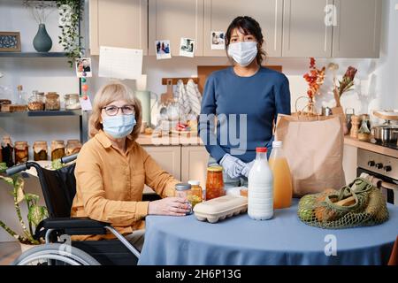 Ritratto di donna anziana con sedia a rotelle seduta al tavolo con prodotti e guardando la macchina fotografica con l'operatore sanitario in piedi vicino, indossano le maschere Foto Stock