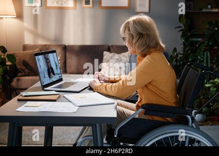 Donna anziana che utilizza una sedia a rotelle seduta al tavolo e parla online con il collega sul laptop mentre lavora a casa Foto Stock