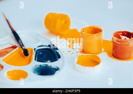 Su un tavolo bianco ci sono vasi con gouache luminoso di colori diversi, e accanto ad esso si trova una tavolozza e un pennello colorato con vernice. Il processo Foto Stock