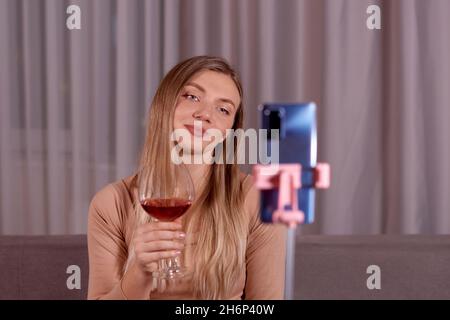 Giovane bella donna che ha alla videochiamata notturna, con smartphone, seduta sul divano, bere vino rosso. Concetto di relazione a lunga distanza Foto Stock